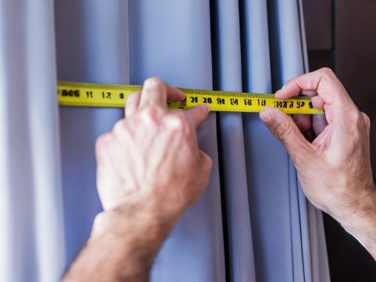 Une main mesure un rideau avec un mètre ruban jaune contre un mur.